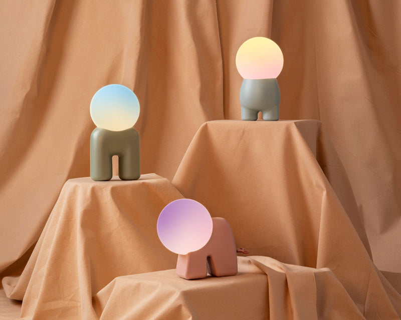 Three small sculptural table lamps shaped like abstract figures with round glowing heads, displayed on draped peach fabric steps against a matching draped backdrop.